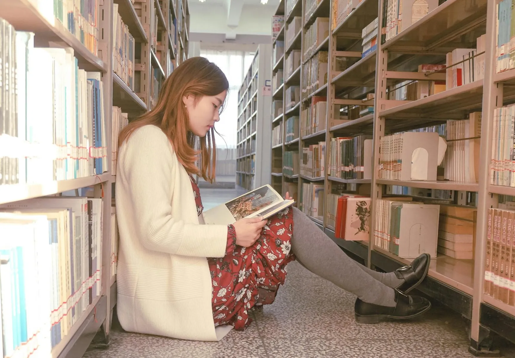 Asian woman sitting and reading book in library aisle surrounded by bookshelves